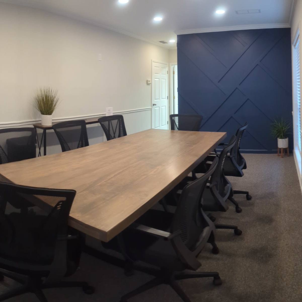 A spacious conference room features a long wooden table surrounded by black mesh chairs, with a door leading outside and a potted plant in the corner.