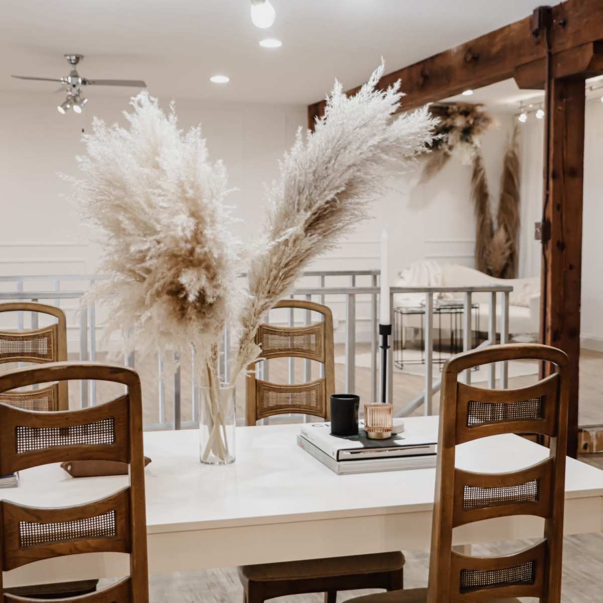 A dining table is set with a vase containing large pampas grass, surrounded by wooden chairs in a well-lit, modern dining area.