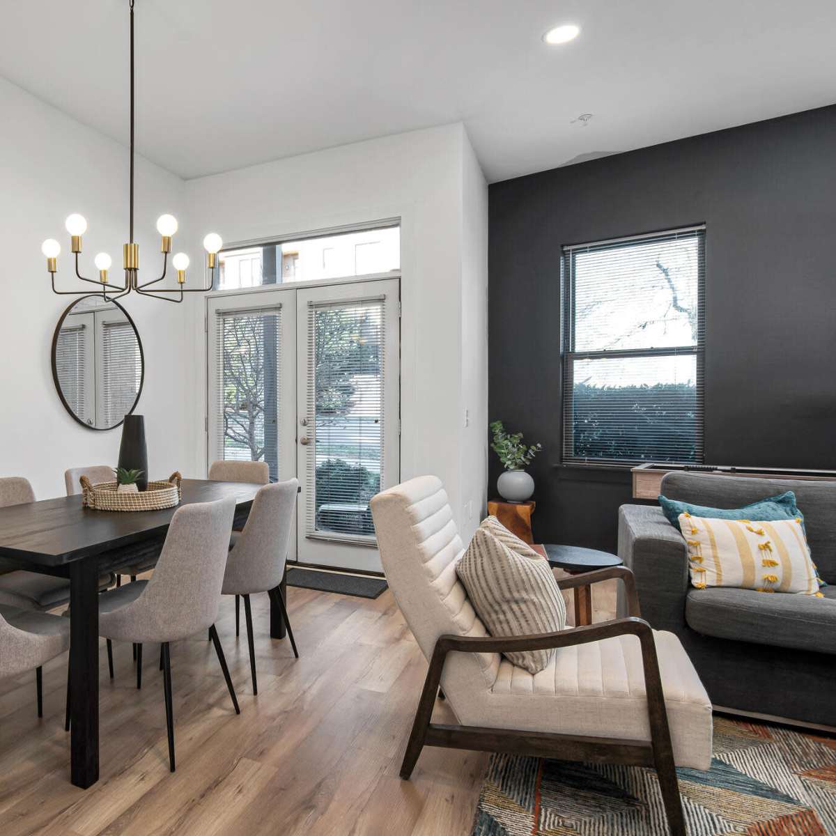 A modern dining area with a black table surrounded by beige chairs, a decorative mirror on the wall, and large windows allowing natural light.