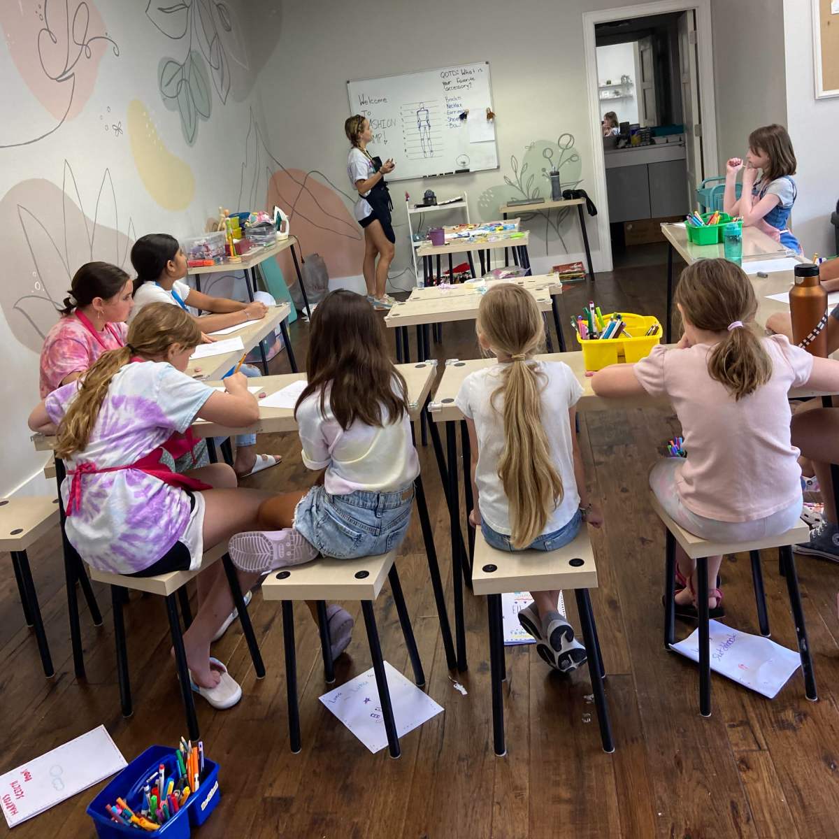 A group of children is seated at small tables in a classroom while an instructor stands at the front, presenting a lesson.
