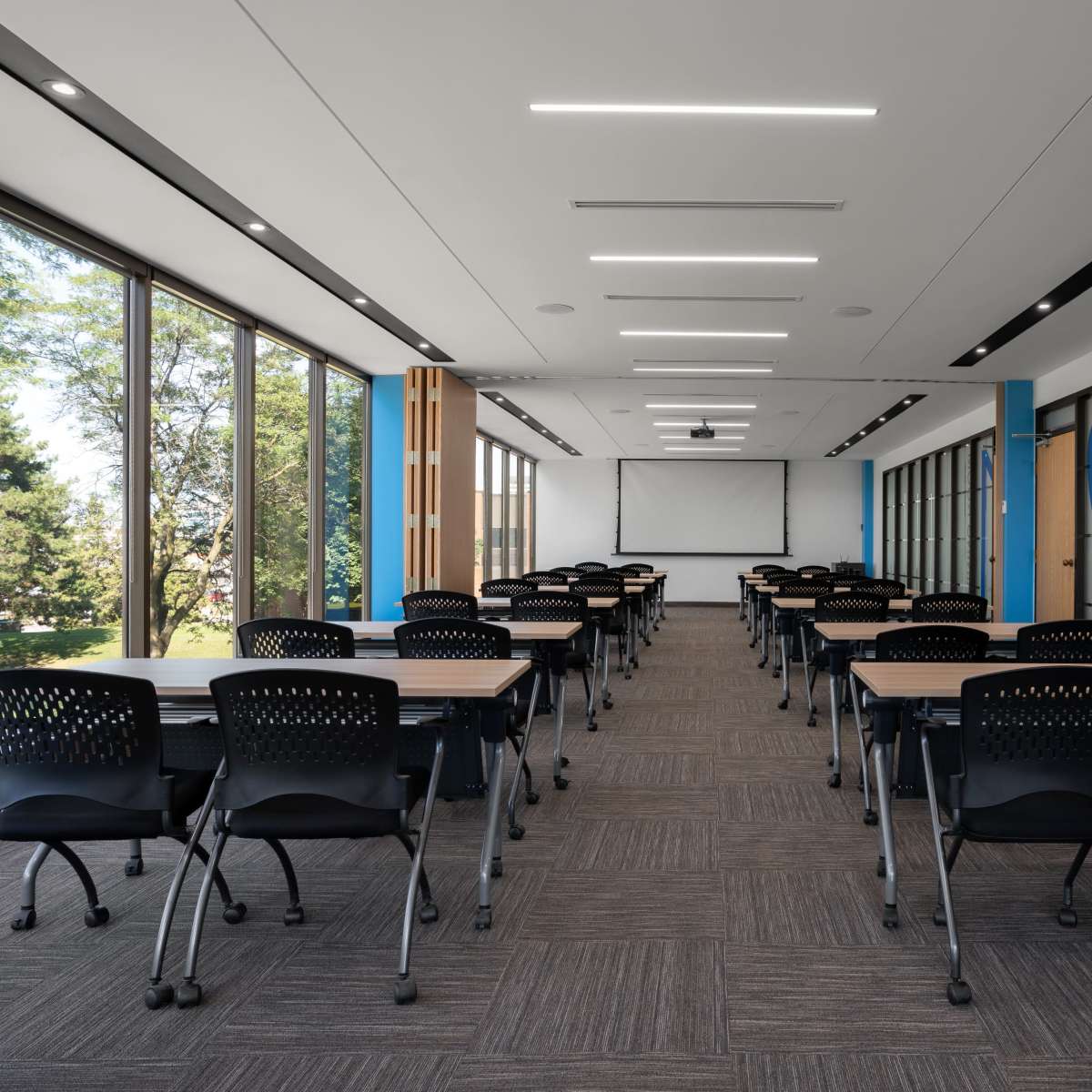A modern classroom features rows of black chairs and tables, large windows allowing natural light, and a projector screen at the front.