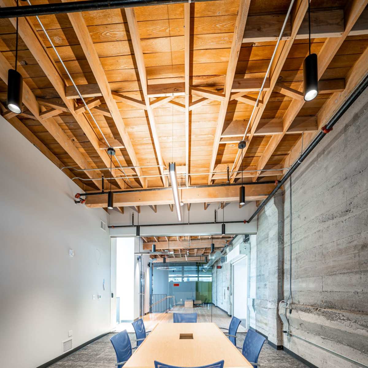A conference room features a long wooden table surrounded by blue chairs, with exposed wooden beams and concrete walls.