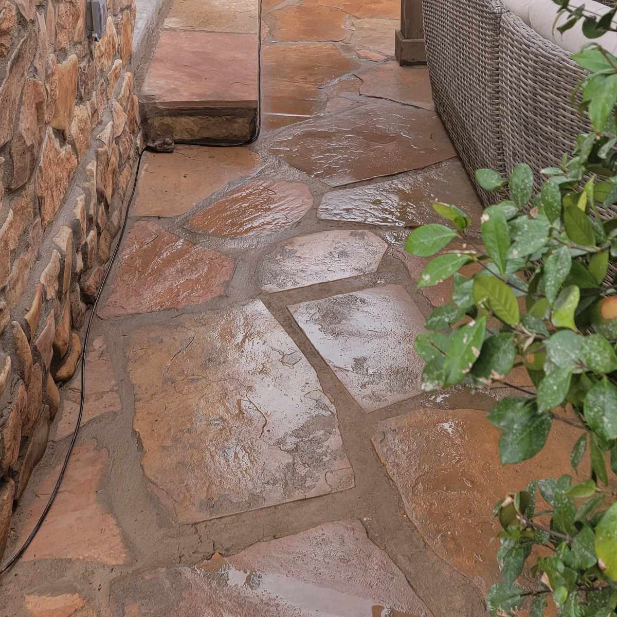 A stone pathway flanked by a textured stone wall and potted plants, leading to a pair of black doors under a covered patio.
