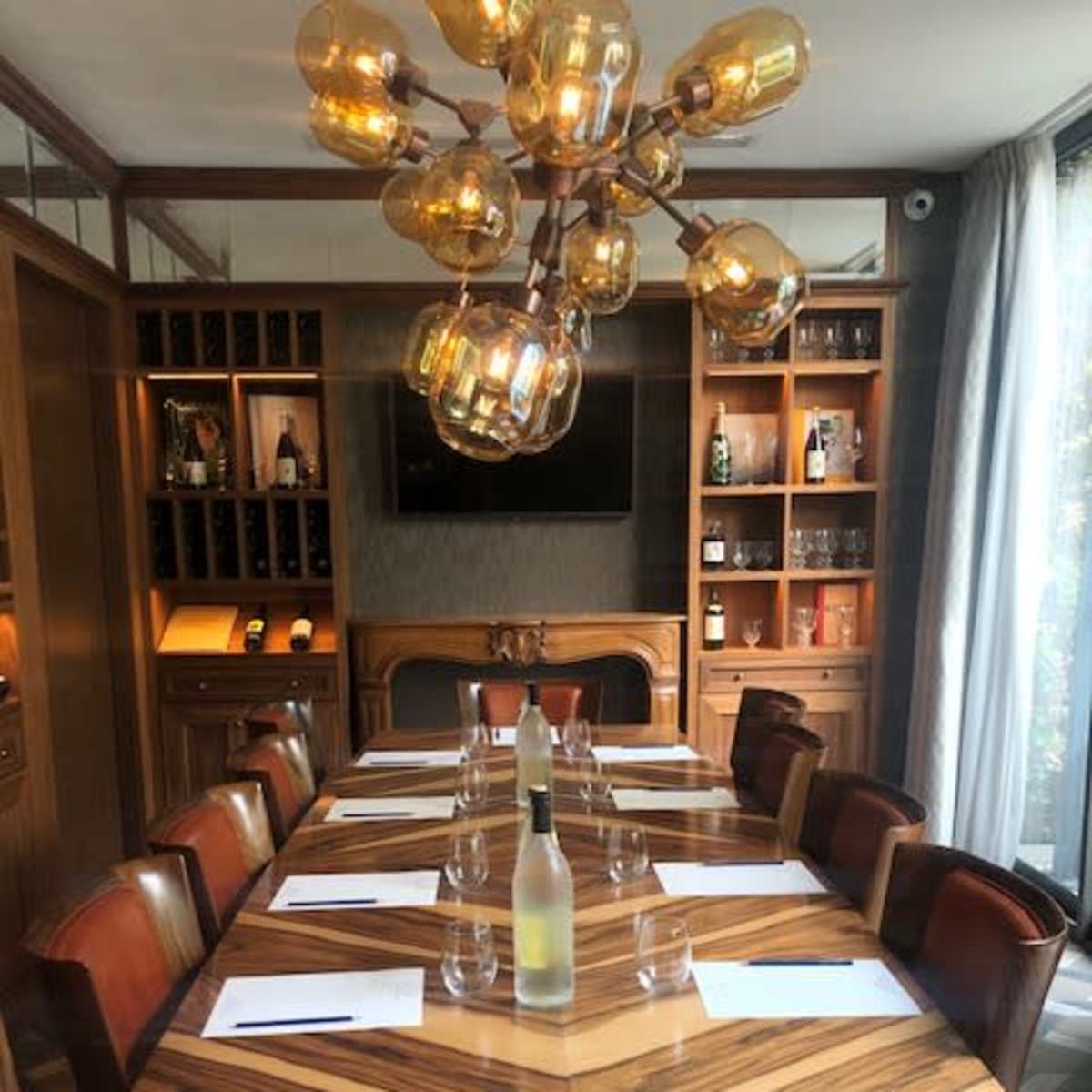 A long wooden conference table is set with glasses and notepads, surrounded by leather chairs, with a modern chandelier overhead and shelves displaying bottles in the background.