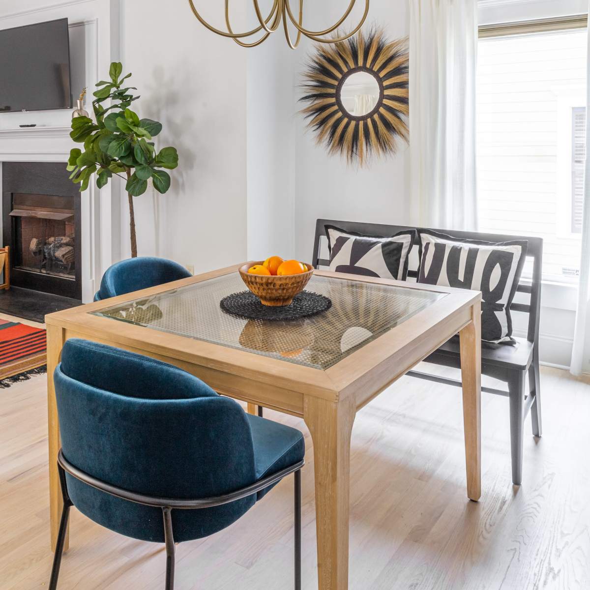 A modern dining area features a wooden table with a glass top, surrounded by blue chairs, accented by a gold chandelier and a decorative mirror on the wall.
