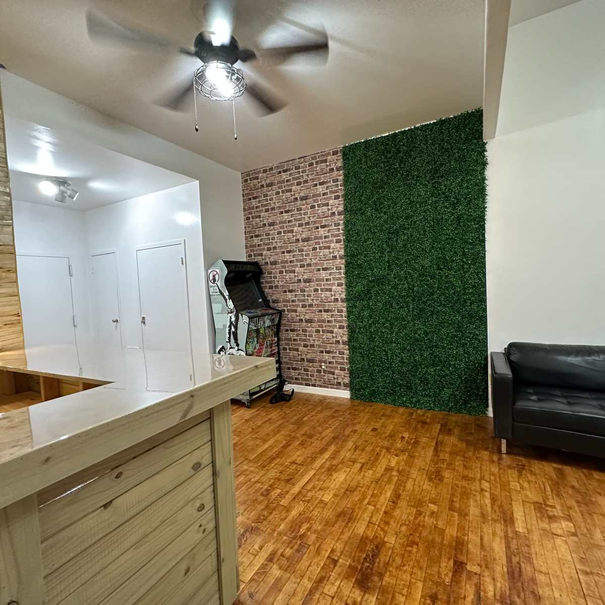 The room features a wooden bar counter, a black leather couch, and a green faux grass wall alongside a brick-patterned wall.