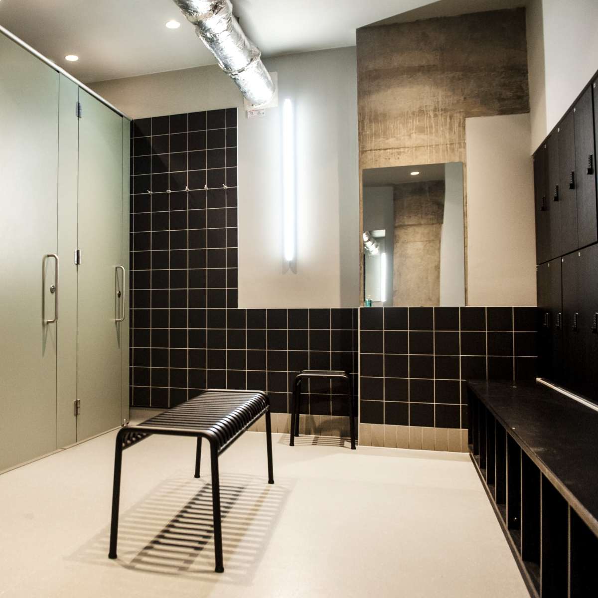A minimalistic changing room with several closed gray lockers and a black bench against a tiled wall.