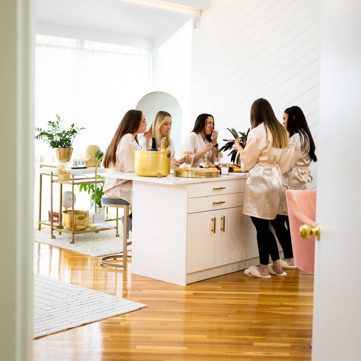 A group of five women in matching robes gathers around a kitchen island, engaging in conversation while preparing food and beverages.