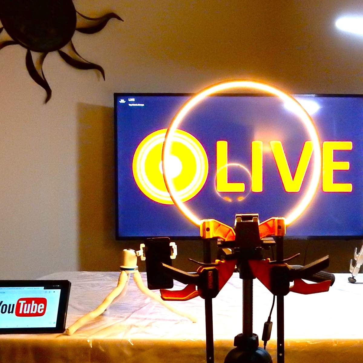 A setup for a live streaming broadcast, featuring a television displaying "LIVE," a ring light, a tripod, and two tablets with the YouTube logo.