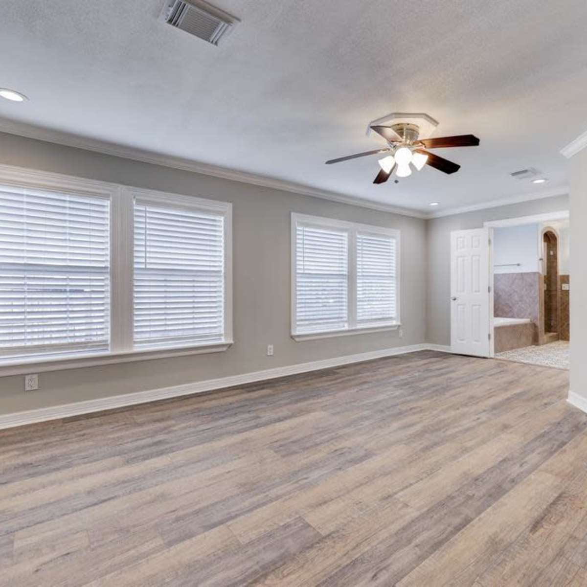 A spacious, light-filled living room with wooden flooring, multiple windows, and a ceiling fan, adjacent to a bathroom area.