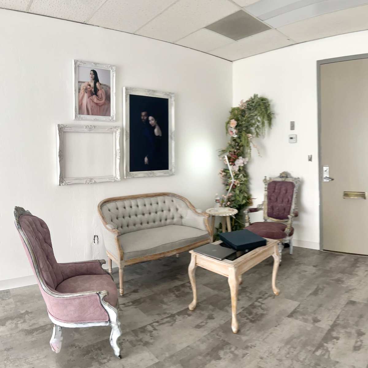 A softly lit waiting area featuring a vintage seating arrangement, including a pink upholstered chair and a cream-colored couch, adorned with floral decorations and framed photographs on the walls.