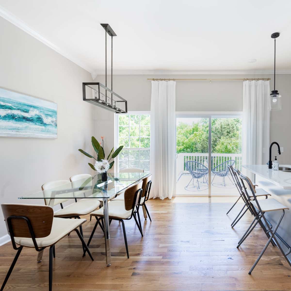 A modern kitchen-dining area features a glass dining table, wooden chairs, and a sliding door that opens to a balcony overlooking greenery.