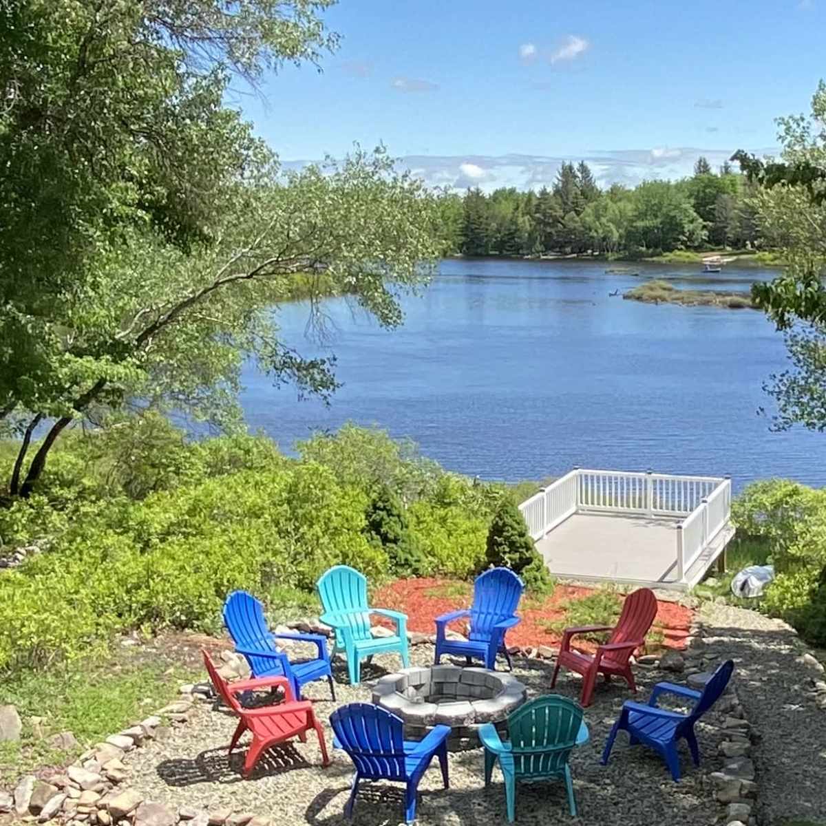 A circular arrangement of colorful chairs surrounds a stone fire pit near a calm river, with green trees and a blue sky in the background.