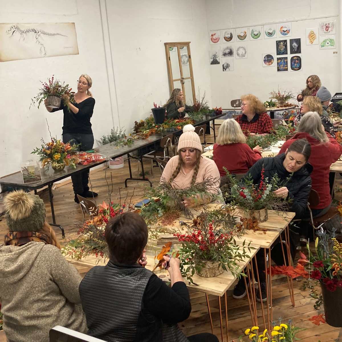 A group of people gathers around tables in a spacious room, engaged in floral arranging with a variety of plants and decorations.