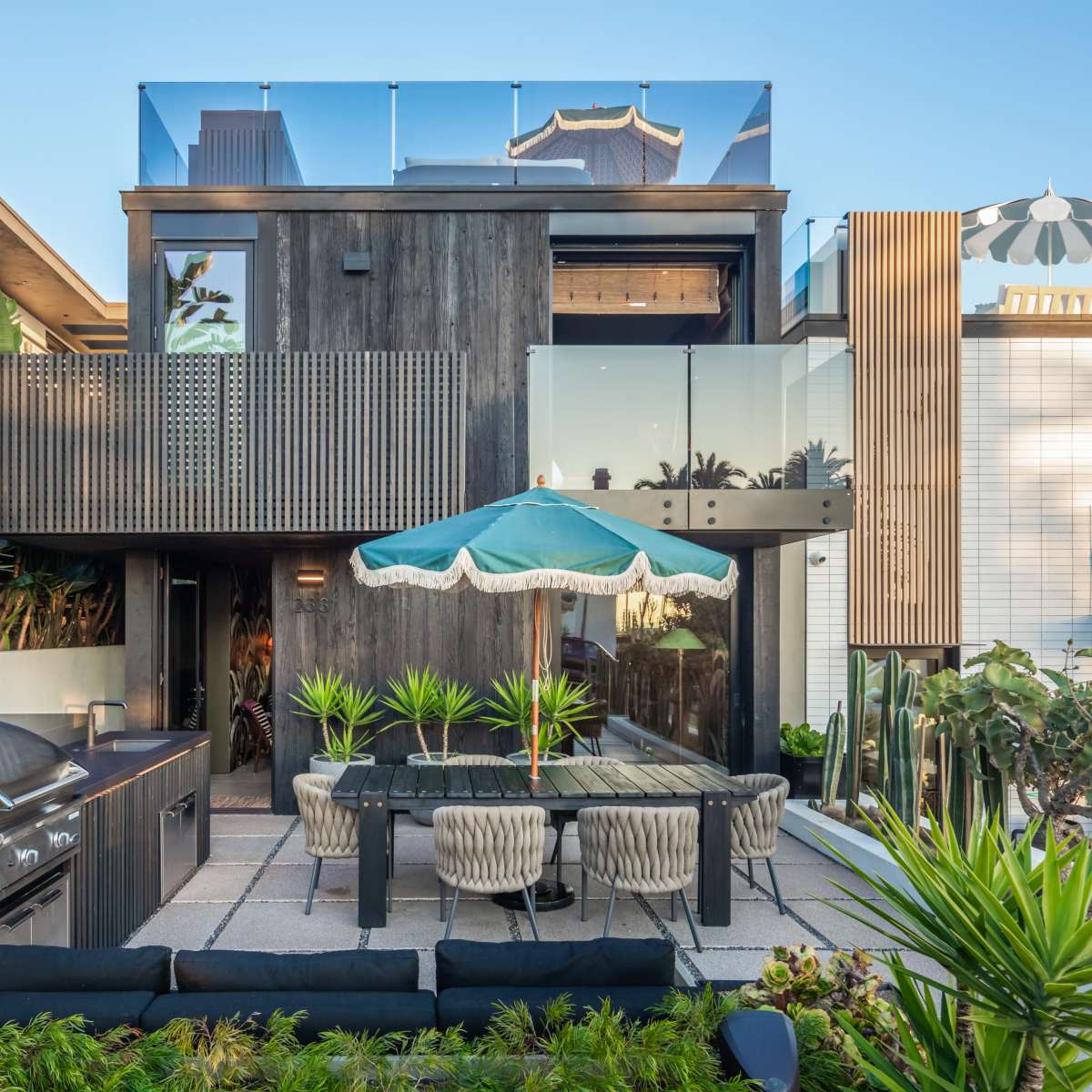 A modern house with wooden exterior features, a glass balcony, an outdoor dining area with a green umbrella, and a grill situated in a landscaped yard.