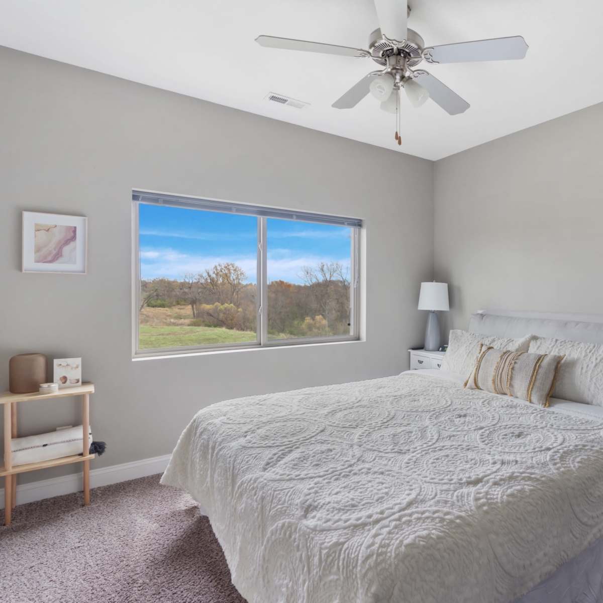 A neatly arranged bedroom featuring a large bed with a white quilt, two bedside tables with lamps, and a window overlooking a grassy landscape.