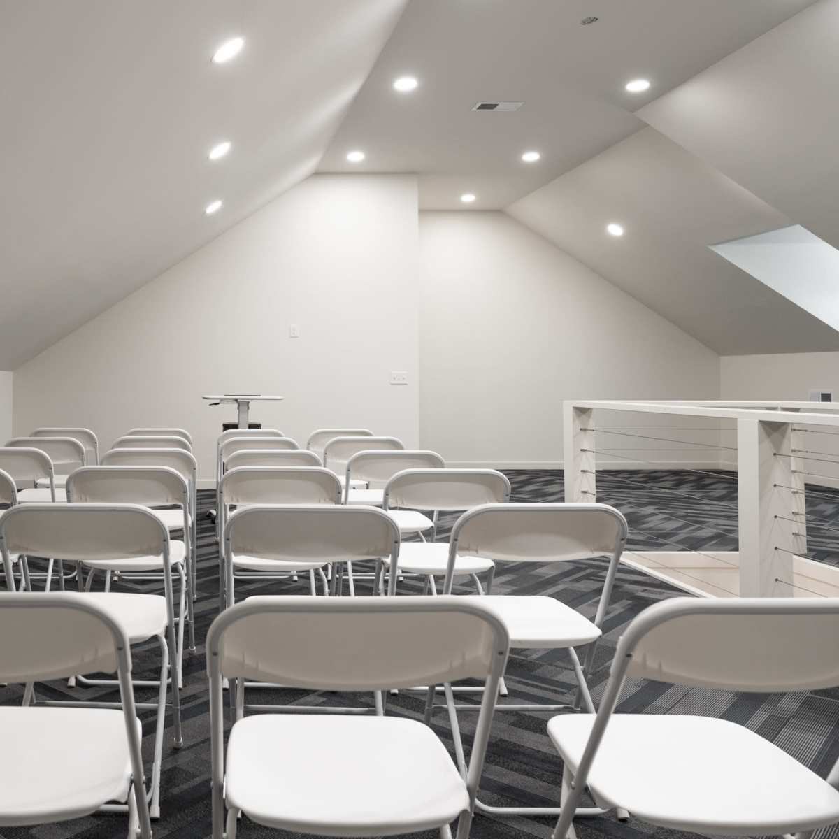 A room with sloped ceilings arranged with several rows of white folding chairs facing a central table.