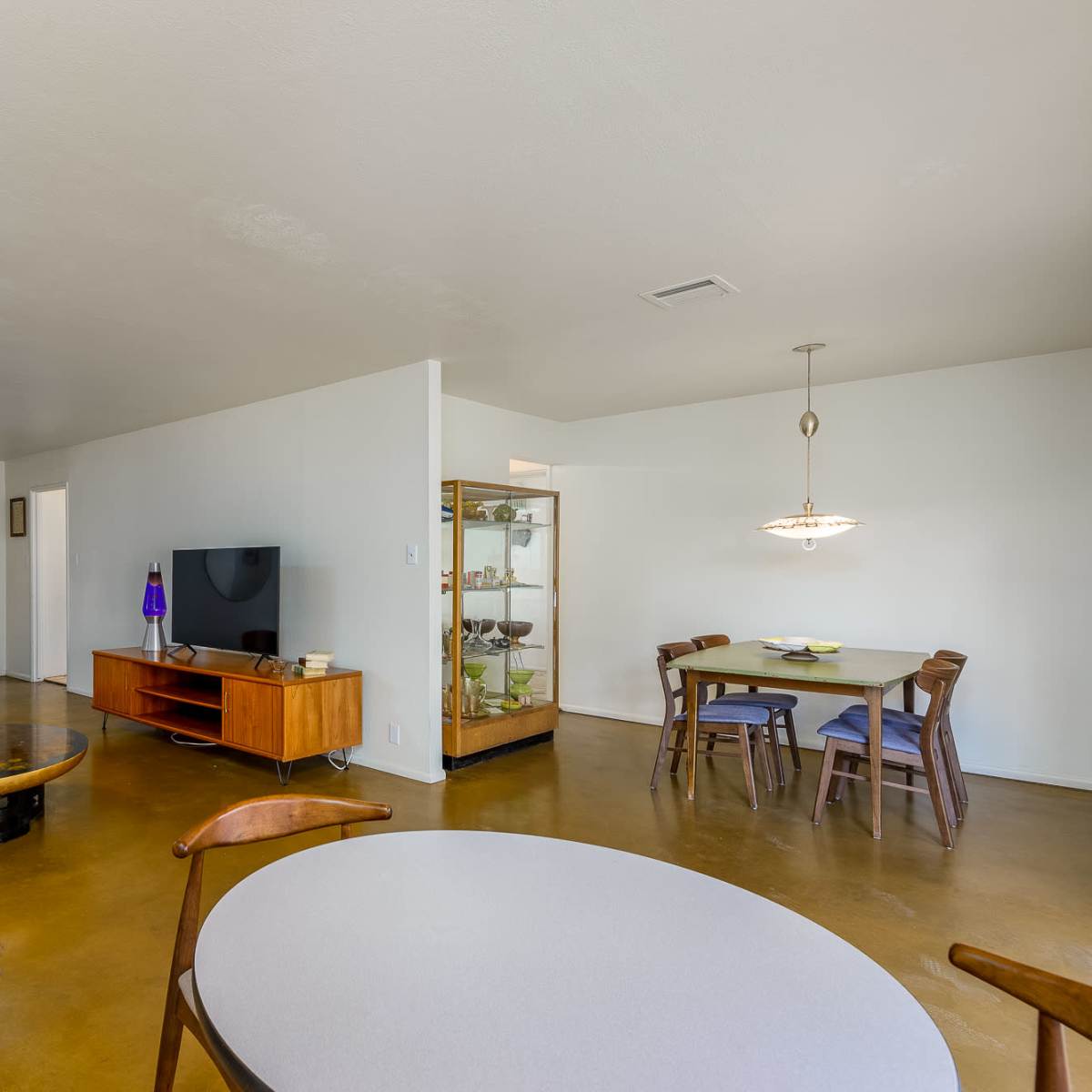 A spacious living area featuring a round dining table, a television on a wooden media console, and a glass display cabinet, with a mix of modern and vintage furniture.
