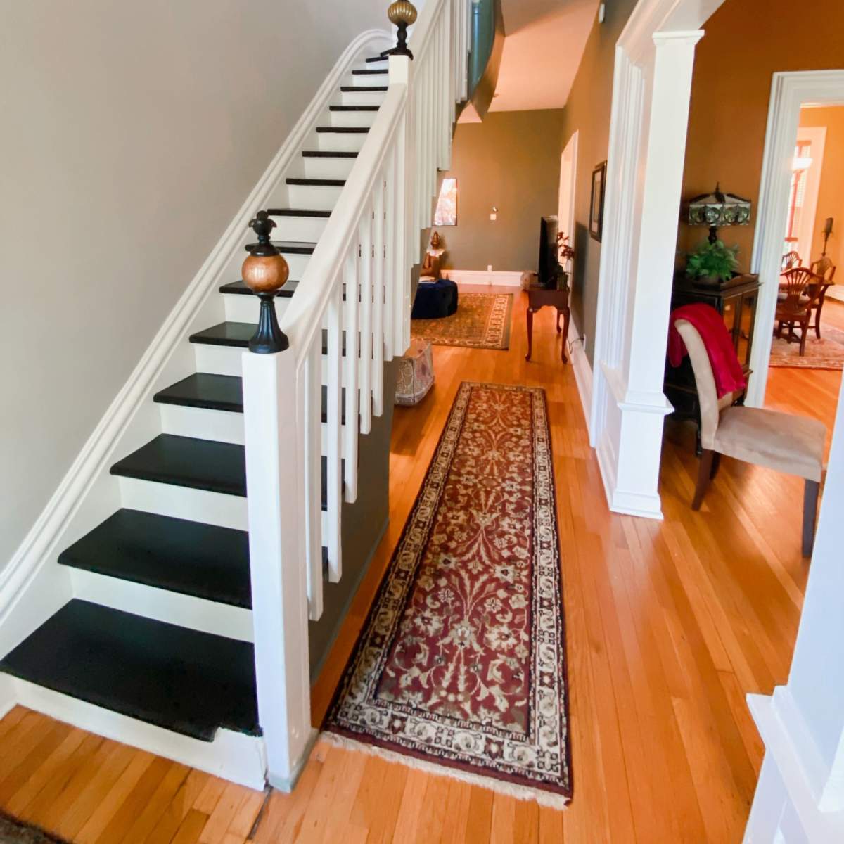 A hallway with a staircase on the left, a patterned runner rug on the wooden floor, and a view into an adjacent room at the end.