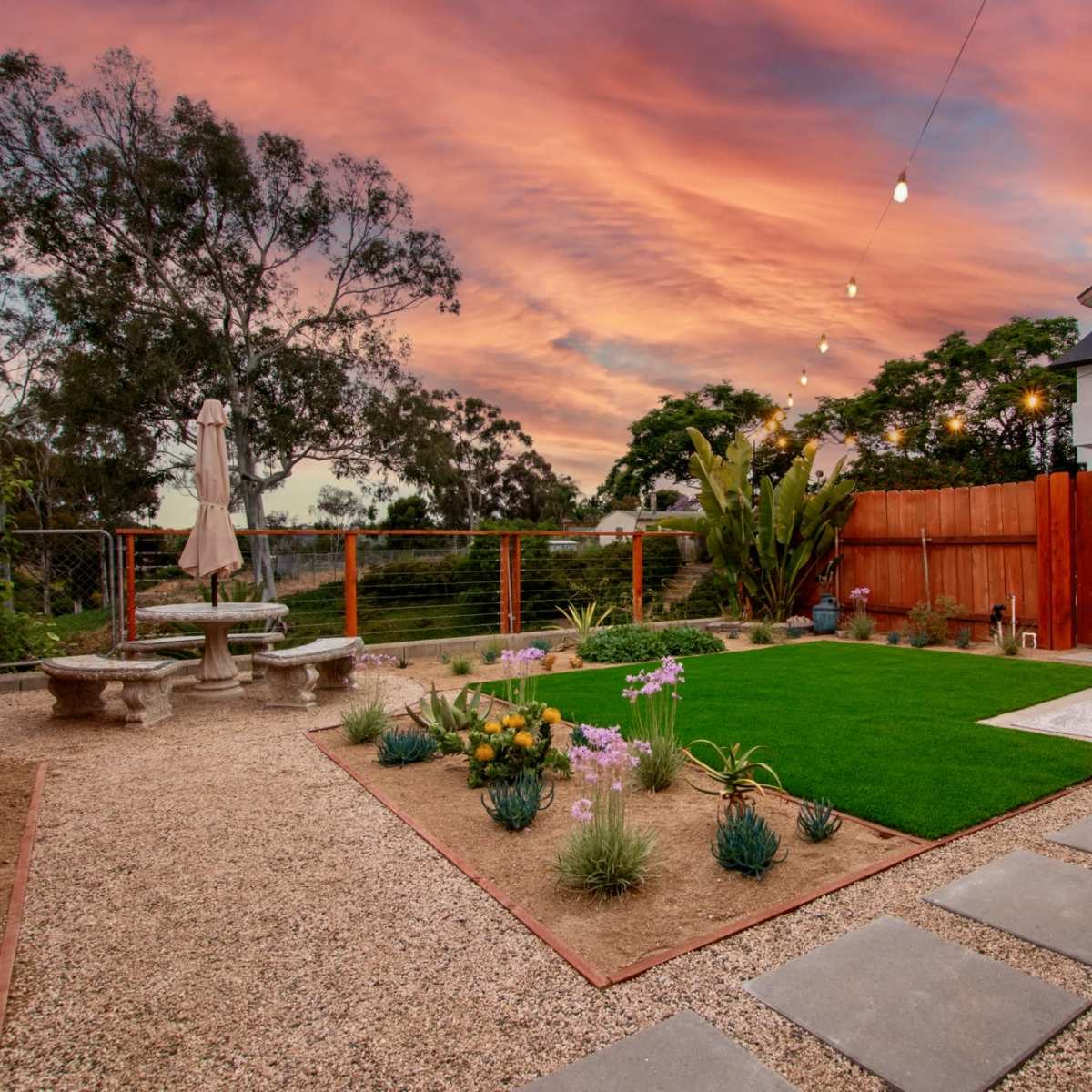 A landscaped backyard with a stone patio, grass area, colorful flowers, and a sunset sky illuminated by string lights.