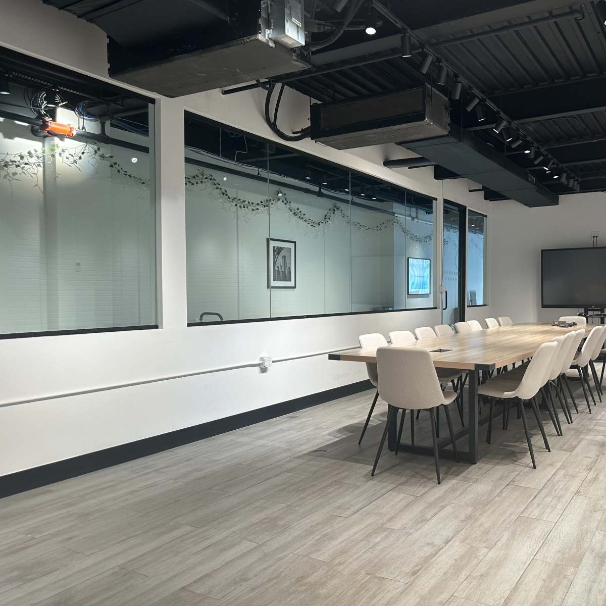 A modern conference room featuring a long wooden table surrounded by twelve chairs, with a large screen and a white brick wall in the background.