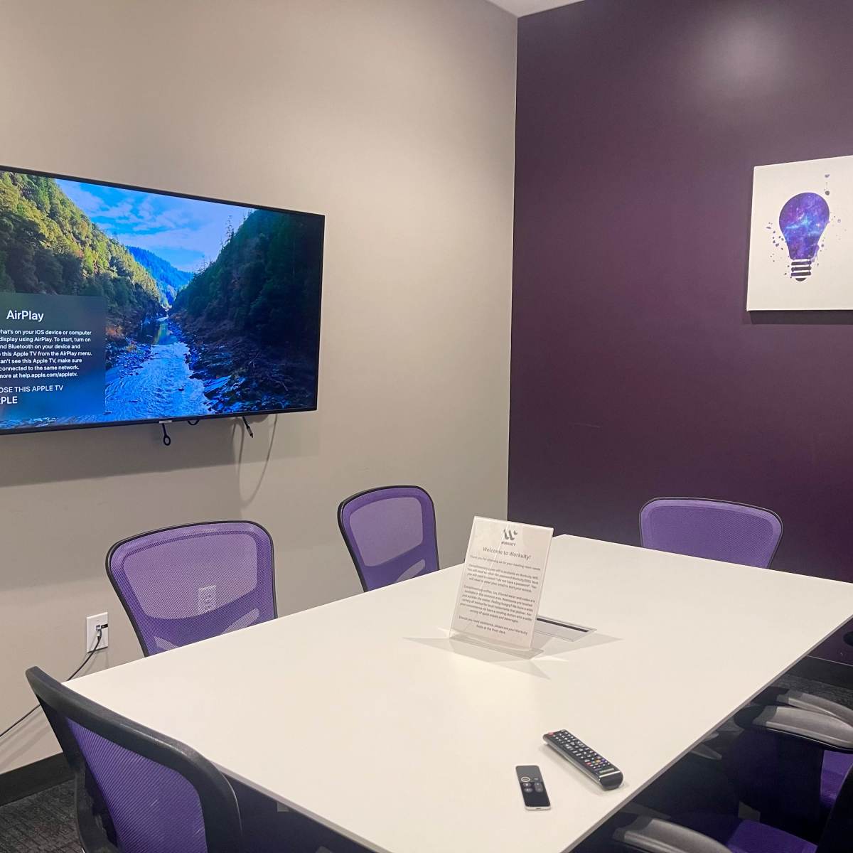 A conference room features a white table surrounded by purple chairs, a large TV displaying a nature scene, and a lightbulb artwork on the wall.