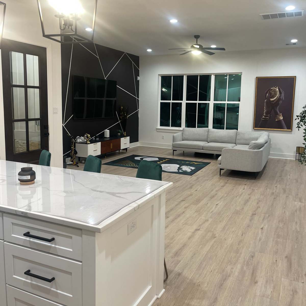 A modern living room with a light-colored sofa, a geometric accent wall, and large windows, viewed from a kitchen with a marble countertop.