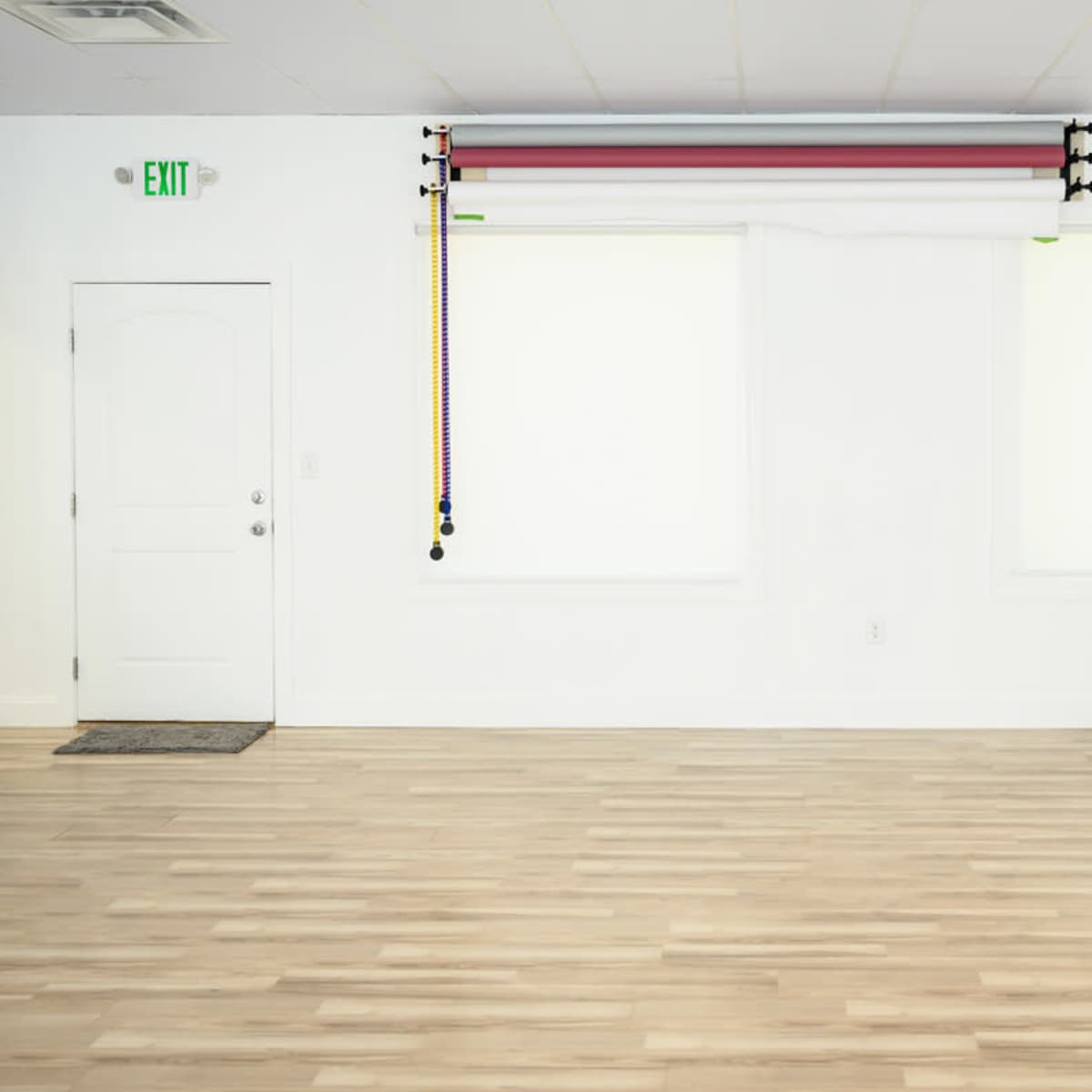 A spacious, empty room with light-colored wooden flooring, a white door, two covered windows, and exercise resistance bands hanging on a wall.