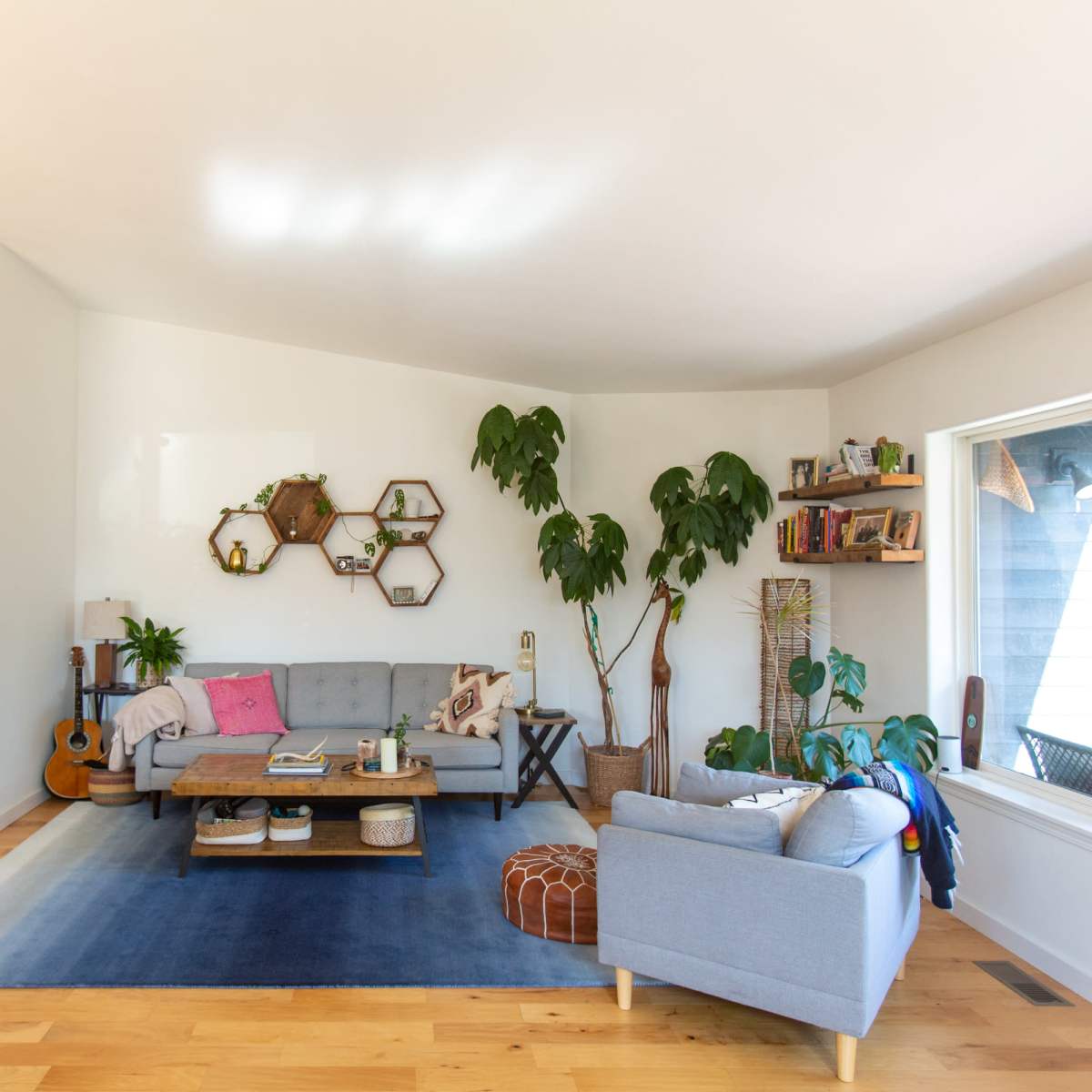 A spacious living room with a gray sofa, plants, a wooden coffee table, and a large window letting in natural light.
