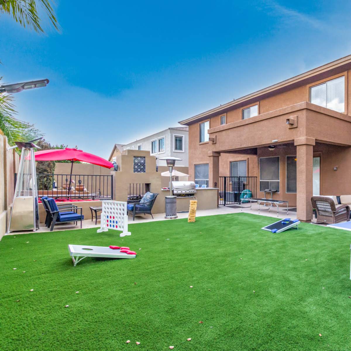 A backyard with artificial grass featuring a ping pong table, cornhole game, and seating areas next to a two-story residential home.