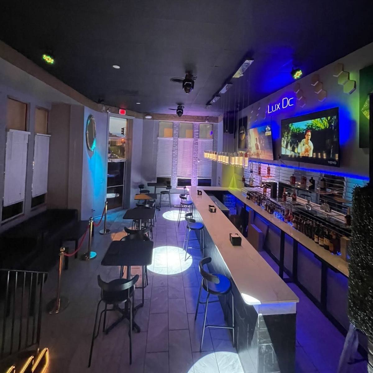 An interior view of a bar with a long counter, high chairs, and ambient lighting, featuring a television mounted on the wall.