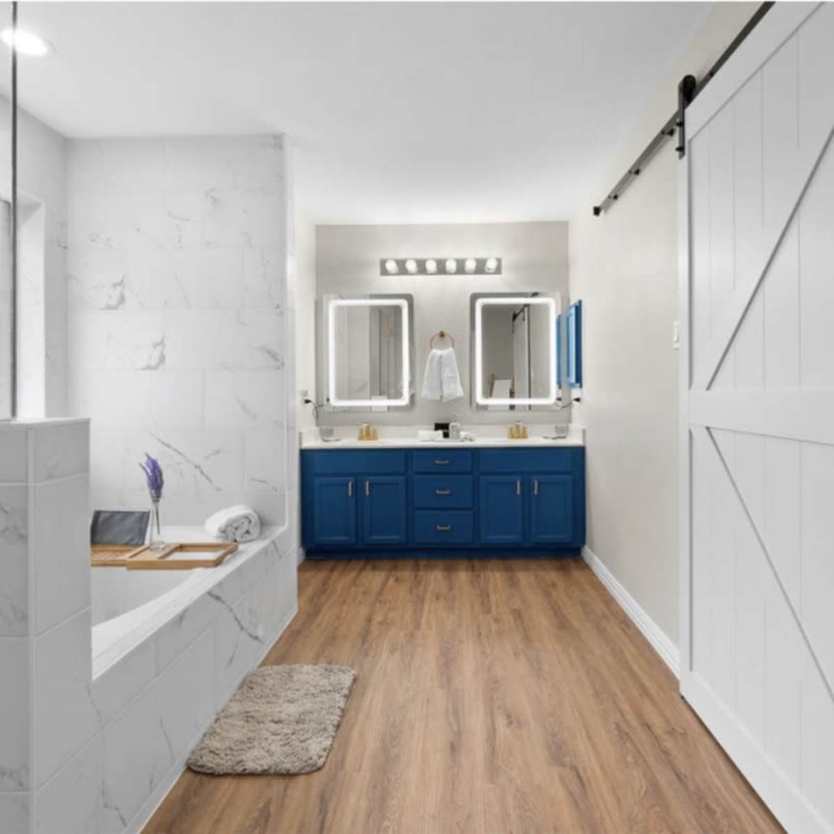 A modern bathroom featuring a freestanding tub, a dual-sink vanity with blue cabinetry, large mirrors with LED lighting, and a sliding barn door.