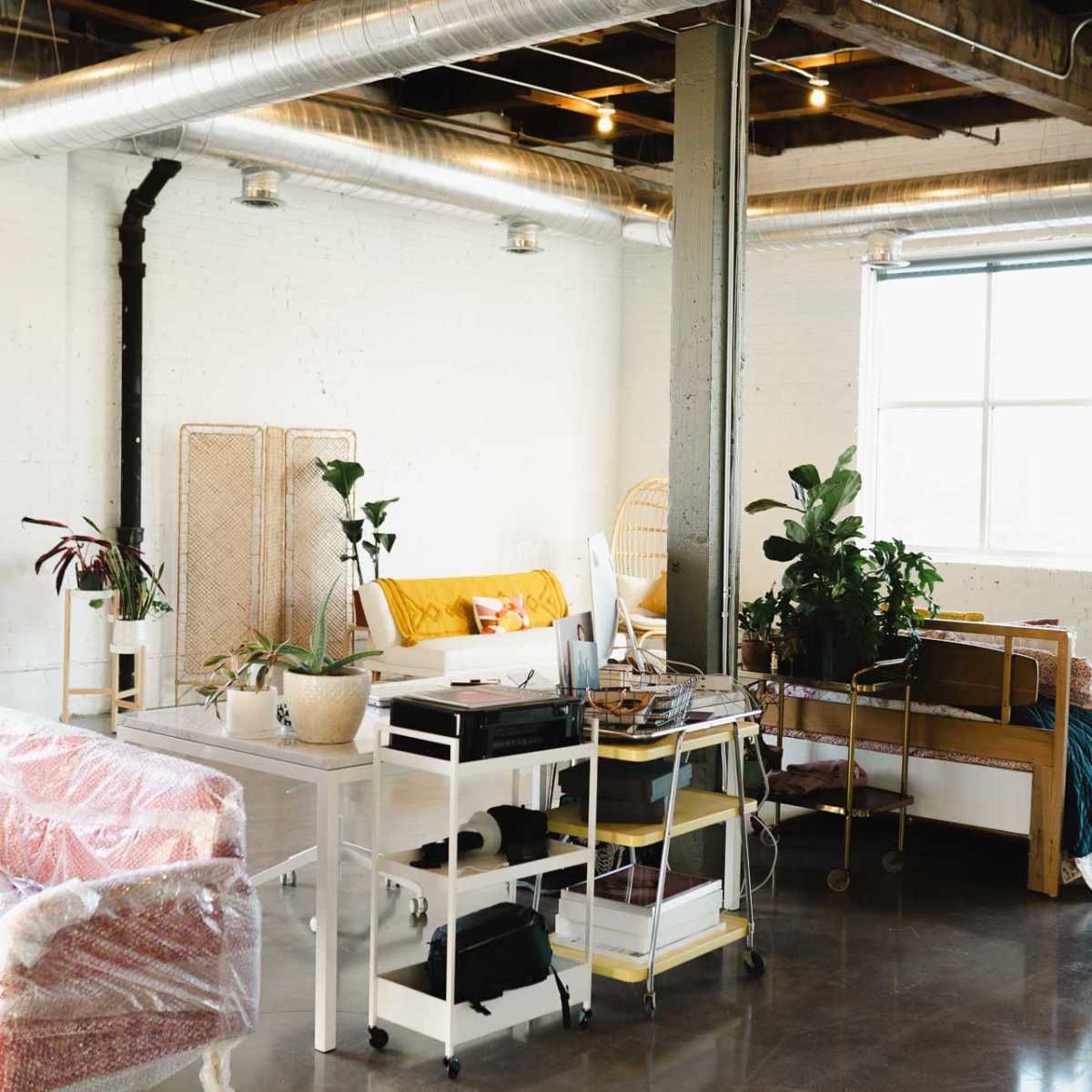 A spacious, modern loft with a seating area featuring a patterned sofa, a plant-filled workspace, and a bed adorned with colorful bedding.