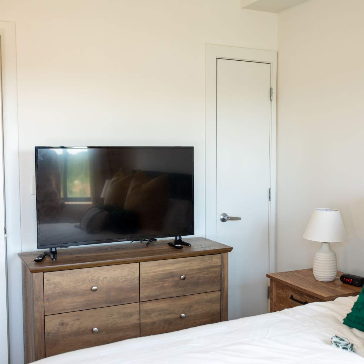 A cozy bedroom with a television on a wooden dresser, a bedside lamp, and a green upholstered headboard.