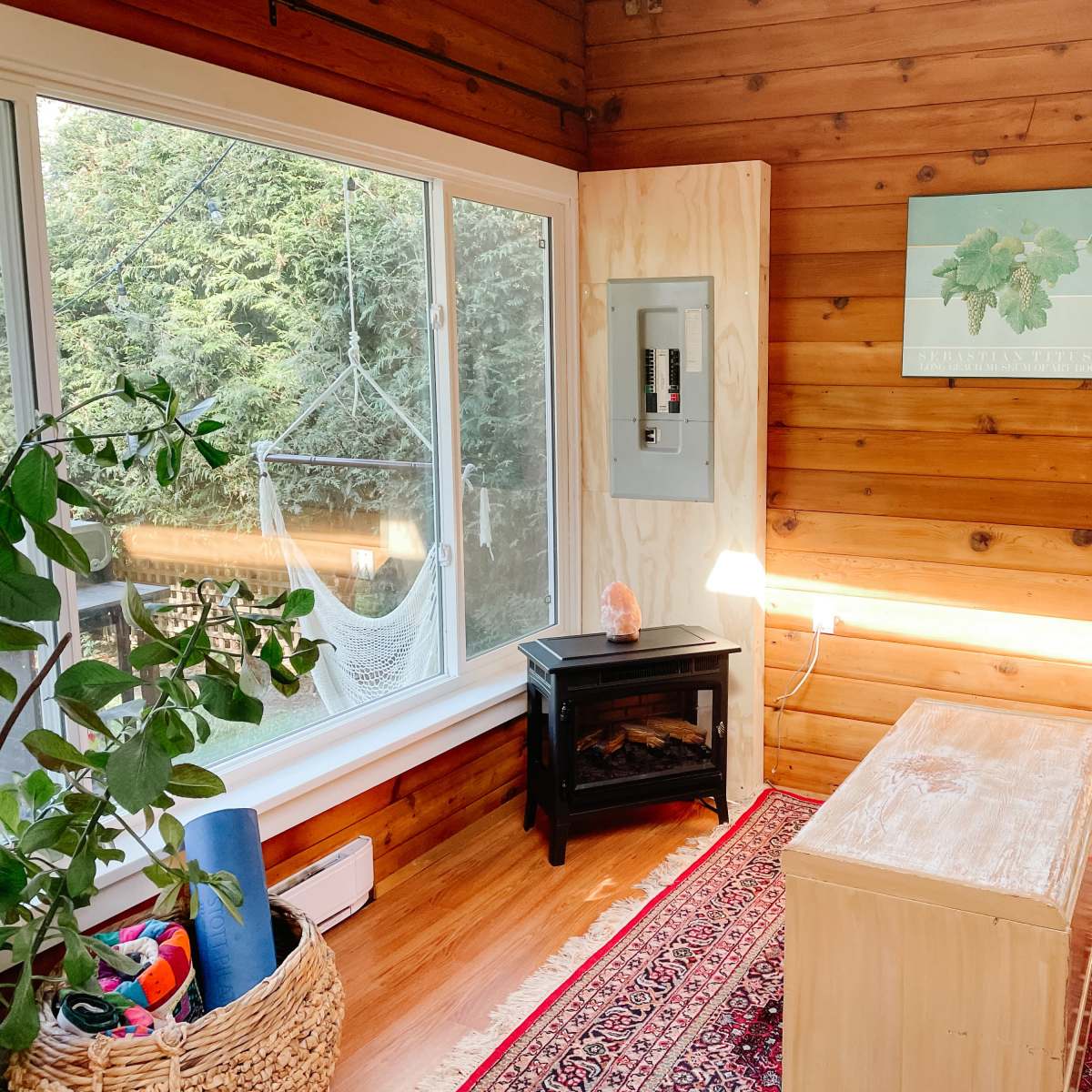 A cozy living space with wooden walls, a small electric fireplace, a coffee table, and a large window overlooking a hammock outside.