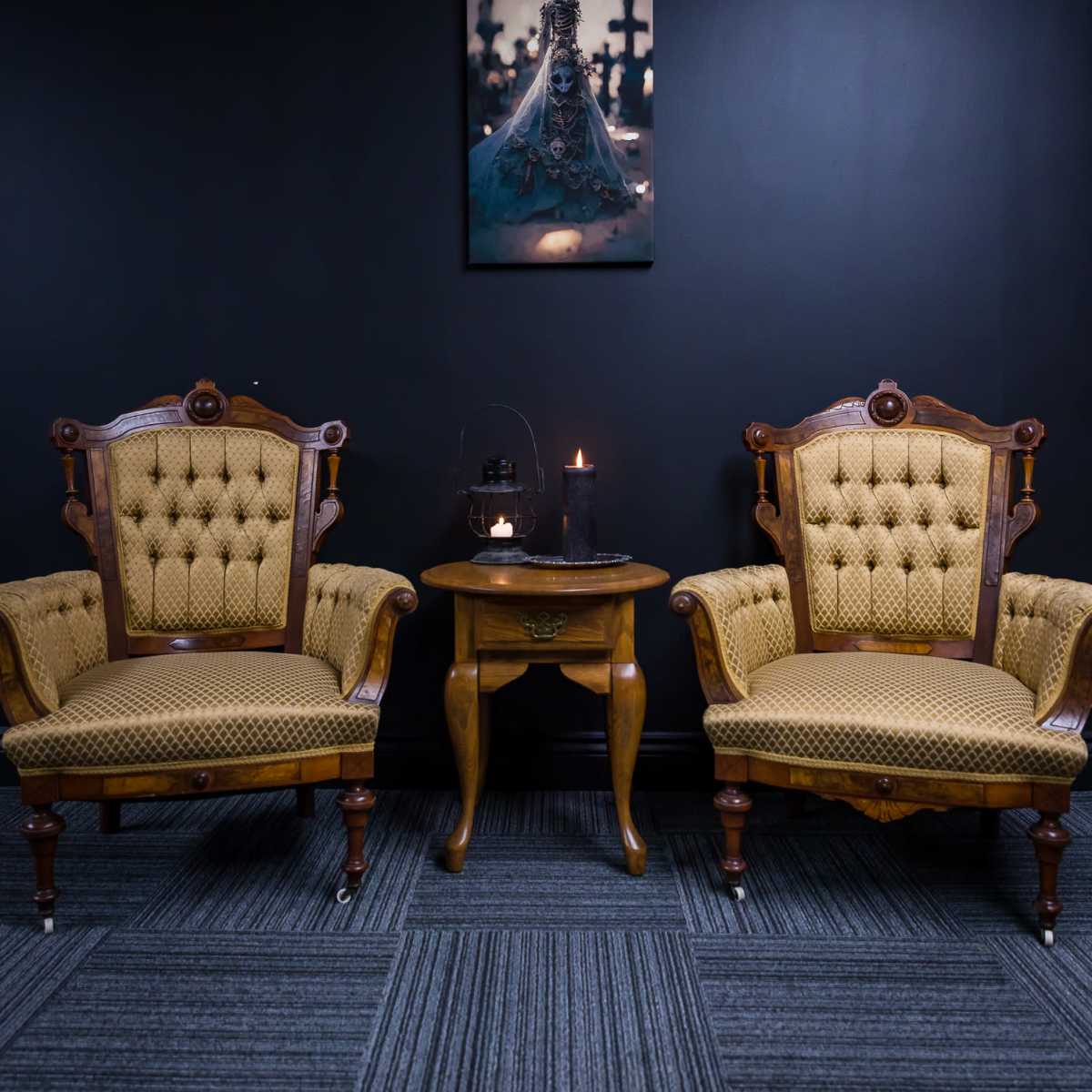 Two vintage tufted chairs flank a small wooden table against a dark wall, with a framed artwork hanging above and candles lit on the table.