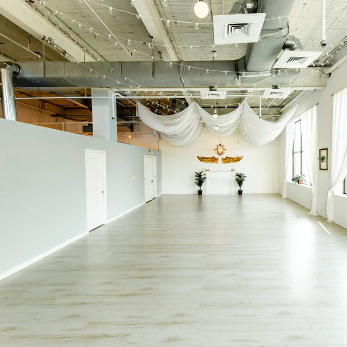 A spacious, well-lit room with large windows, light-colored wooden flooring, and white drapes hanging from the ceiling.