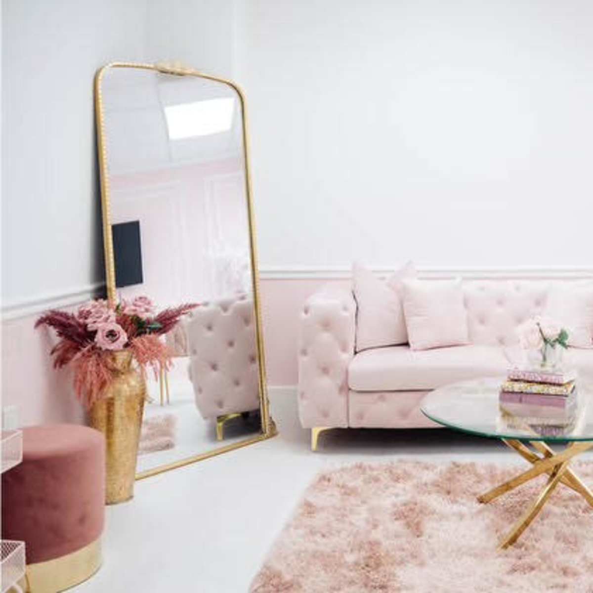 A light pink tufted sofa with pillows, a round glass coffee table, and a large mirror in a bright, softly designed living room.