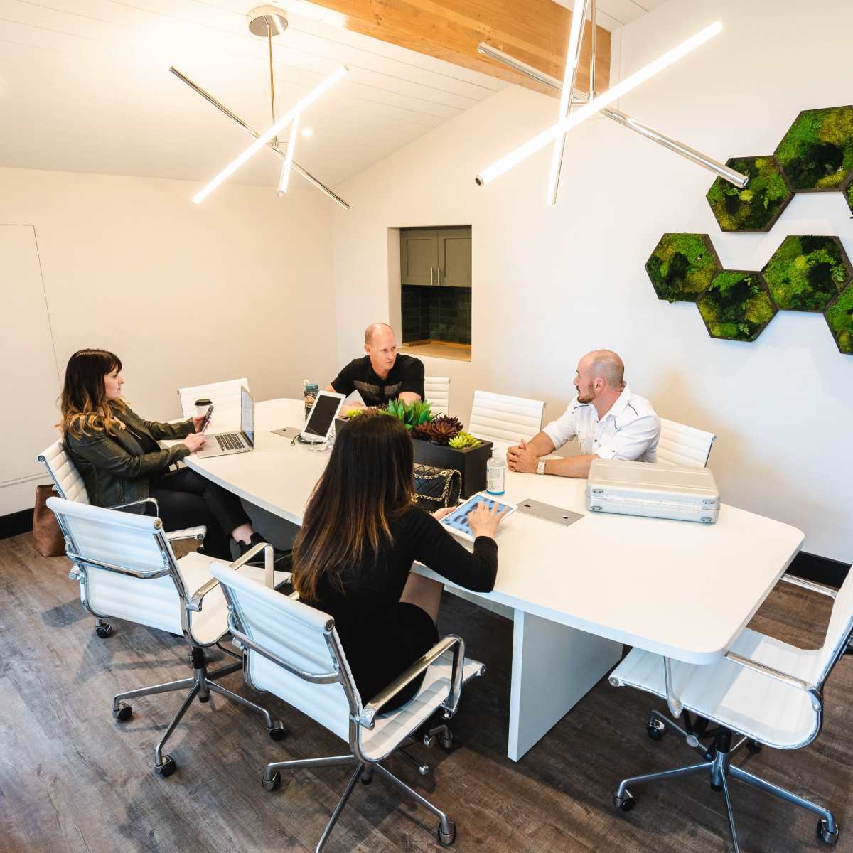 A sleek conference room features a large white table surrounded by four people engaged in discussion, with modern lighting and greenery on the wall.