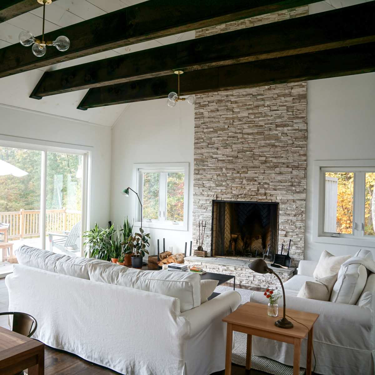 A modern living room with a stone fireplace, large windows, and wooden beams across the ceiling.