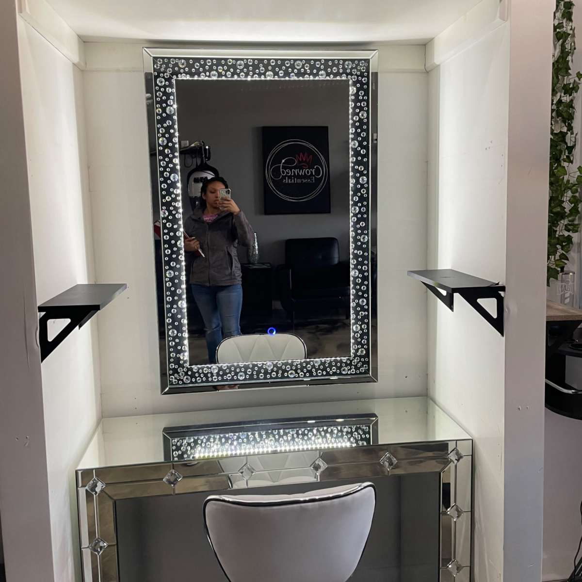 A salon vanity with a large mirrored frame, a white chair, and wall-mounted shelves on either side of the mirror.