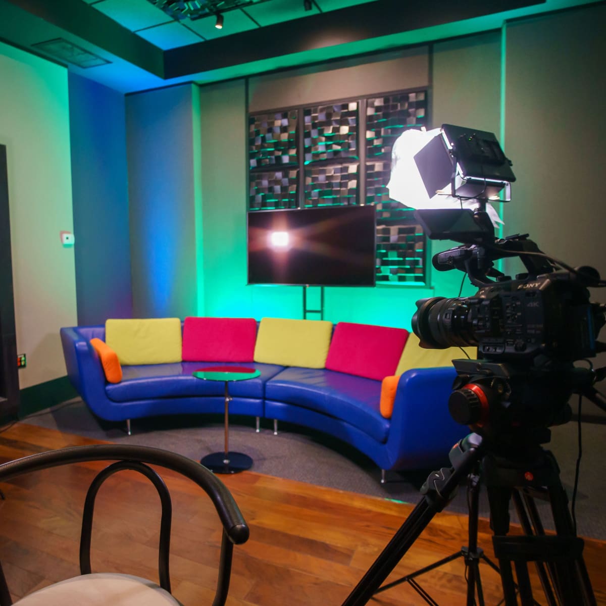 A colorful interview setup with a blue couch, a small circular table, and cameras positioned on either side, illuminated by bright lights.