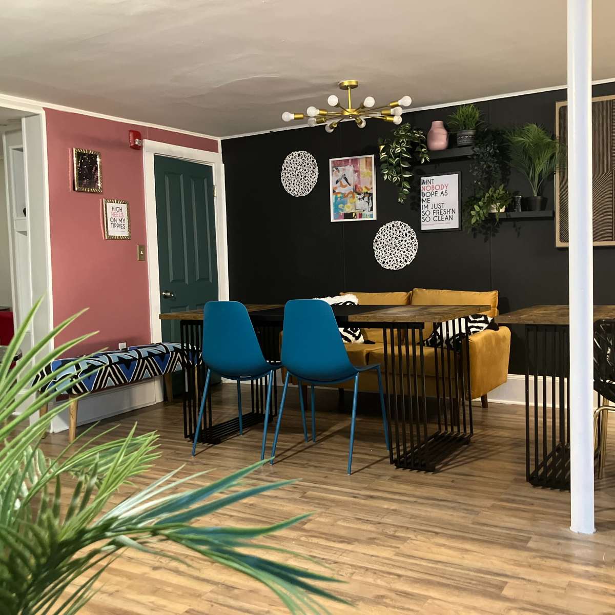 A modern, open living space with a yellow sofa, blue chairs, and tables surrounded by green plants and decorative wall art.