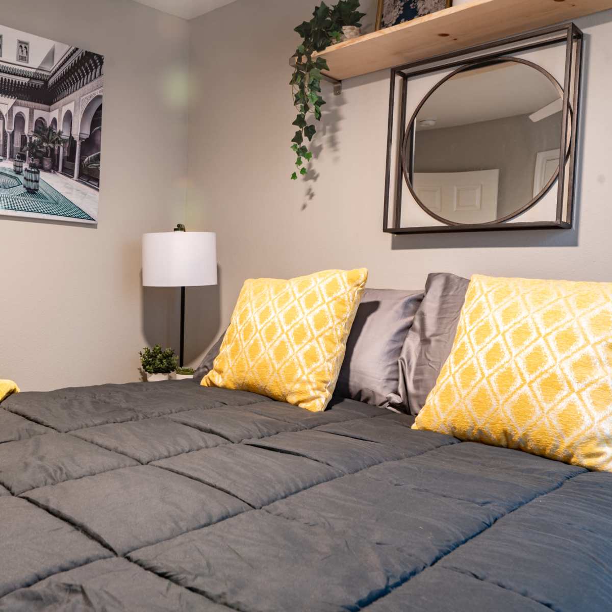 A neatly made bed with gray bedding and yellow accent pillows is positioned beneath a decorative mirror and a wall art piece featuring an architectural courtyard scene.