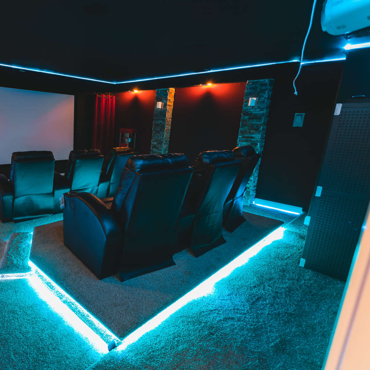 A home theater room with leather recliners, a large screen, and blue LED lighting along the edges of the carpet.