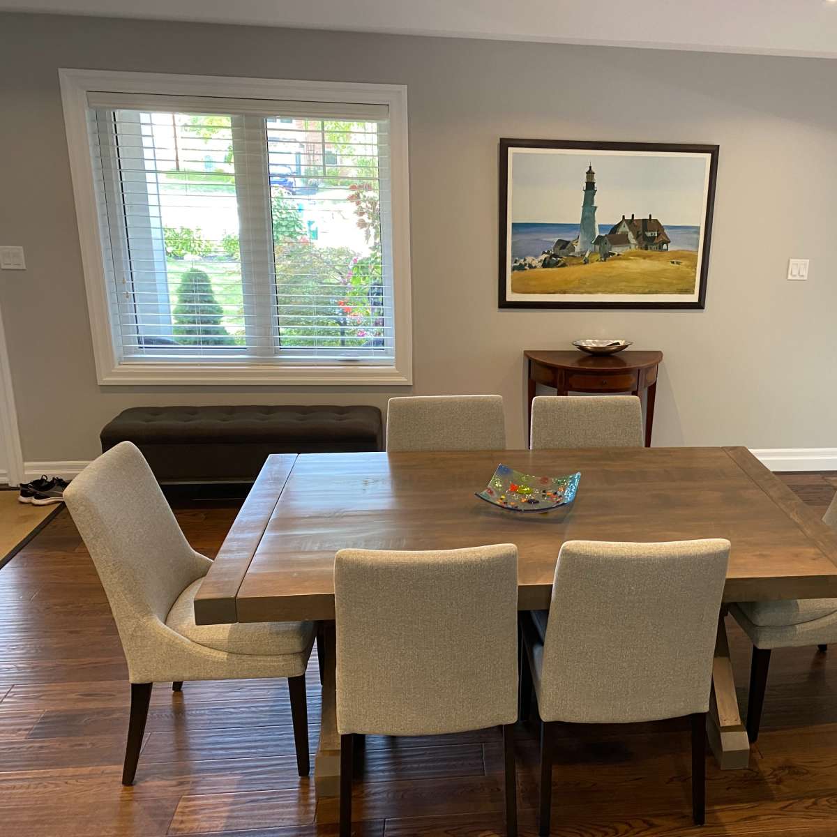 A wooden dining table surrounded by four upholstered chairs is positioned in a well-lit room, with a painting of a lighthouse on the wall and a console table in the background.