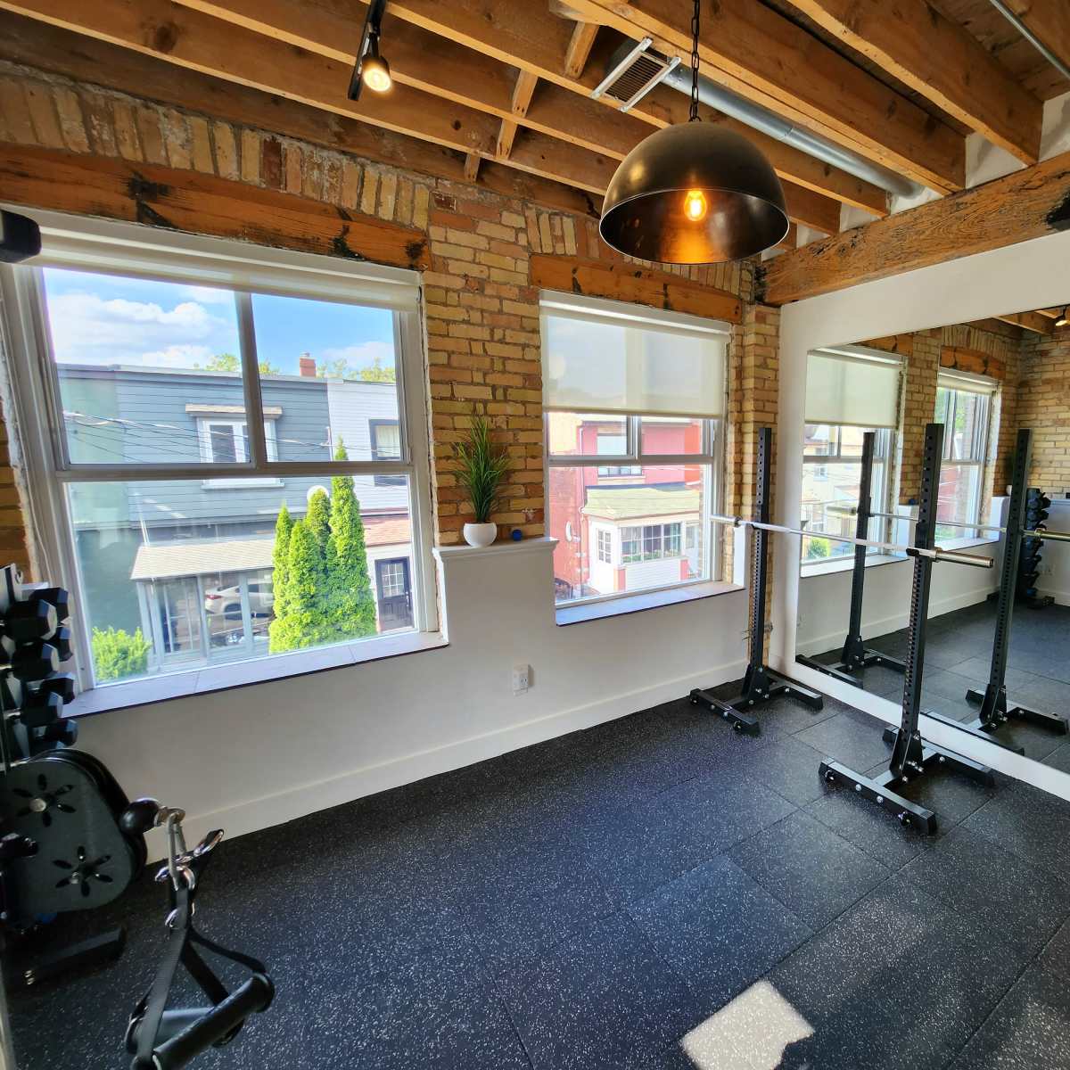 A bright gym with large windows, exposed brick walls, and a mirror, featuring weightlifting equipment and a rubber floor.