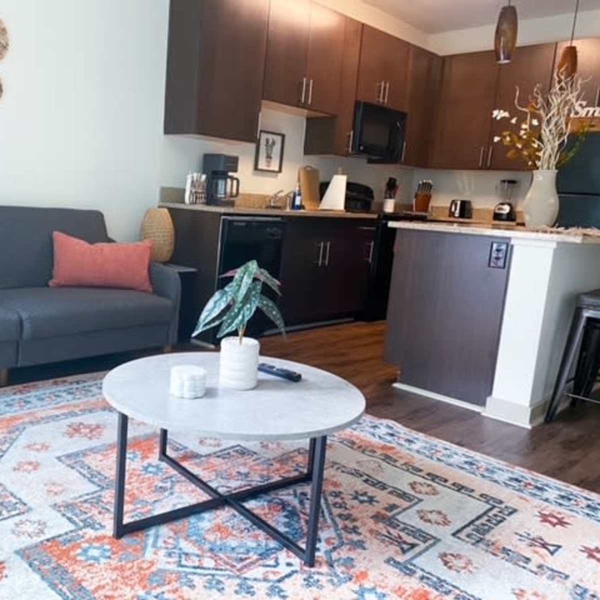 A cozy living room features a gray sofa with pink cushions, a marble-top coffee table, and a colorful area rug, adjacent to a modern kitchen with dark cabinetry.
