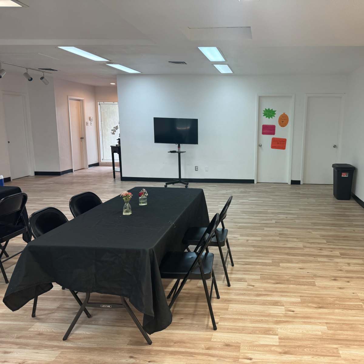 A spacious room with light wood flooring, featuring two tables covered with black tablecloths and several chairs arranged nearby, along with a television mounted on a stand and a trash bin.