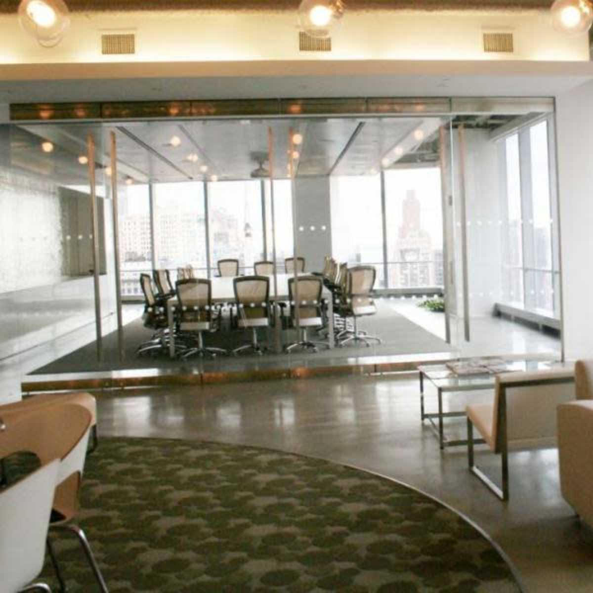 A modern office space features a glass-walled conference room with a large table and chairs, adjacent to a seating area with a couch.